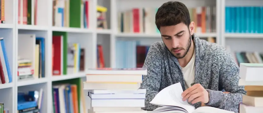 En ung man sitter vid ett skrivbord med en stapel av böcker i ett bibliotek